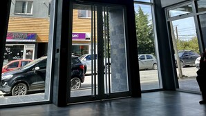 Moderni rakennuksen sisäänkäynti, jossa on suuret lasiovet, näkymä ulkona parkkeerattuihin autoihin ja kattoon ripustetut valaisimet.