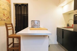 A small kitchen with a white countertop, a wooden chair, a cutting board, a decorative tile, and a black cabinet with a sink.