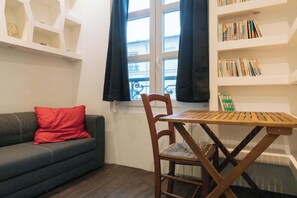 A room with a grey sofa, a wooden table, a chair, a bookshelf, and a window with black curtains.