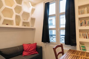 A room with a grey sofa, a red pillow, a wooden table, a bookshelf, and a window with black curtains. - Perfect Studio Getaway for Lovers & Solo Explorers (Paris)