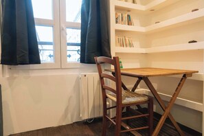 A room with a wooden table and chair, a radiator, a window with curtains, and a bookshelf.