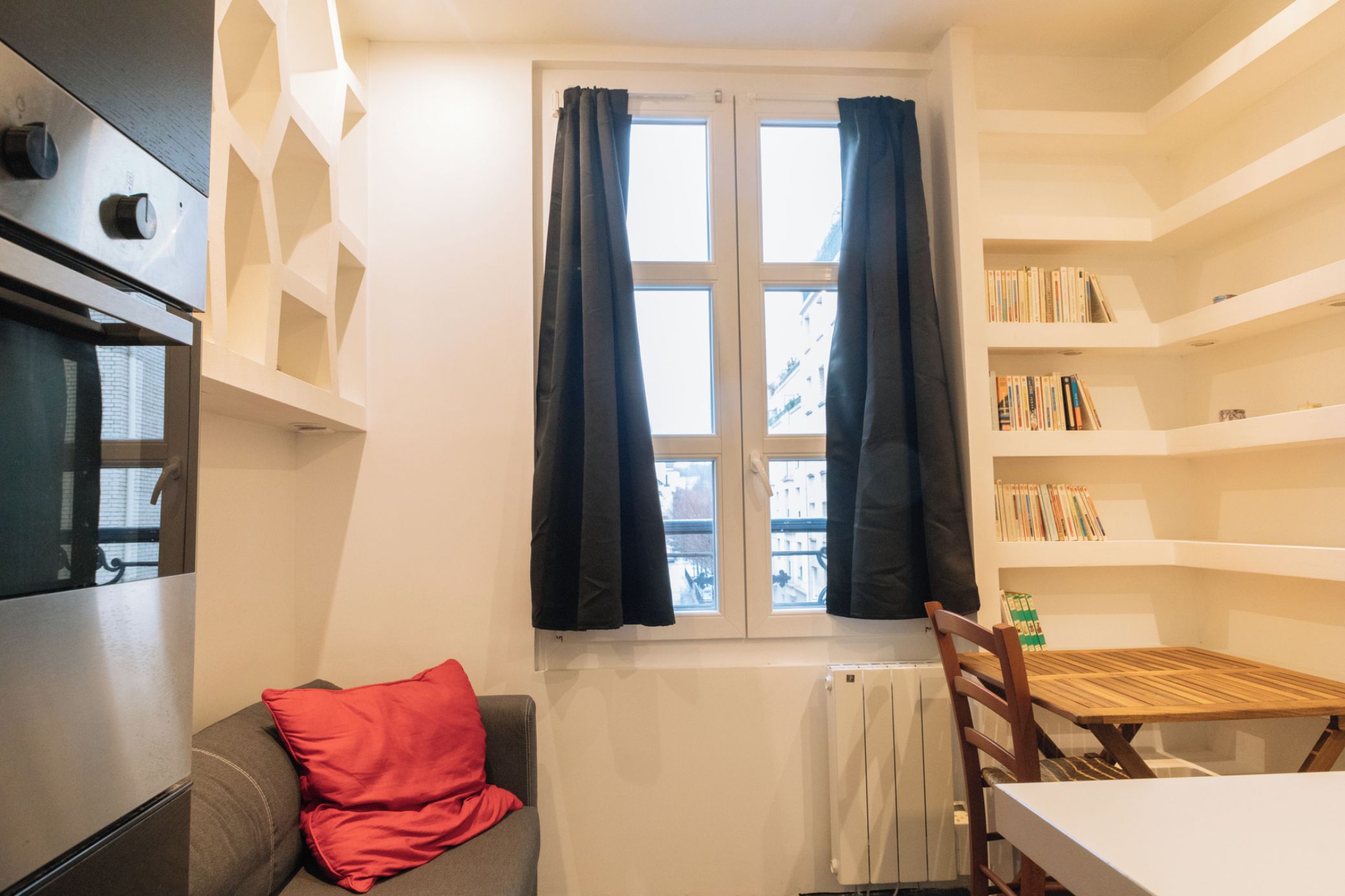 A compact kitchen with a window, a radiator, a bookshelf, and a dining area.