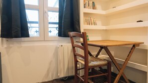 Una habitación con una silla y una mesa de madera, un radiador, una ventana con cortinas y una estantería llena de libros.