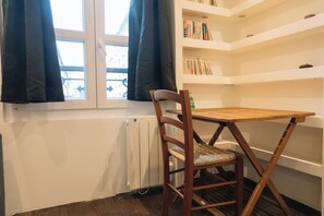 A room with a wooden chair and table, a radiator, a window with curtains, and a bookshelf with books. - Perfect Studio Getaway for Lovers & Solo Explorers (Paris)