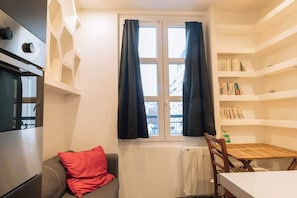 A compact room with a kitchenette, a window with curtains, a bookshelf, a red pillow on a sofa, and a dining table with chairs. - Perfect Studio Getaway for Lovers & Solo Explorers (Paris)