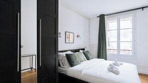 Een net opgemaakt bed met witte lakens en groene kussens, een nachtkastje en een raam met gordijnen.