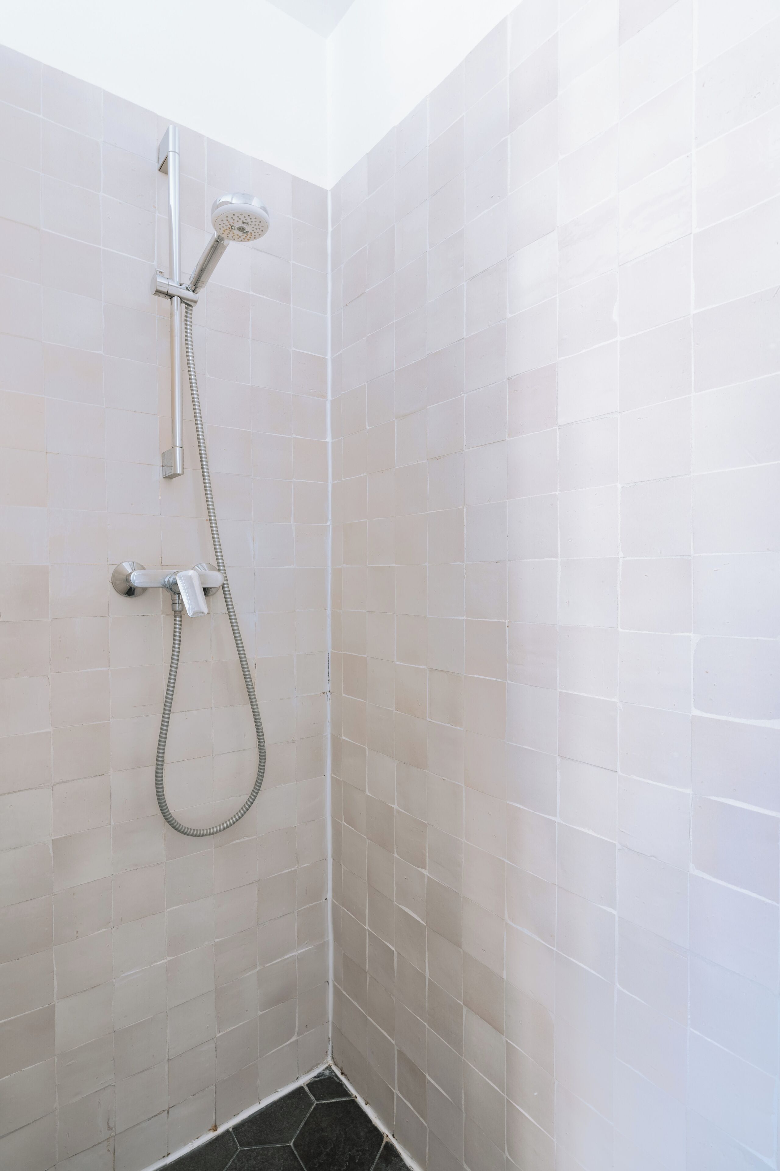 Eine Dusche mit Handbrause und fest installierter Duschbrause, geflieste Wände und ein dunkler Boden.