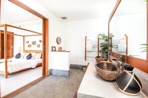 A modern hotel room with a wooden bed, a small desk, a large mirror, and a stone basin. - Modern 3BR Villa Center Seminyak Enclosed Living (Canggu)
