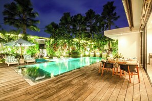 A wooden deck with a swimming pool, lounge chairs, and a table with chairs. - Mette 3br Villa (Canggu)
