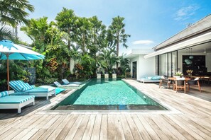 A swimming pool with lounge chairs and umbrellas, surrounded by lush greenery and a wooden deck. - Mette 3br Villa (Canggu)