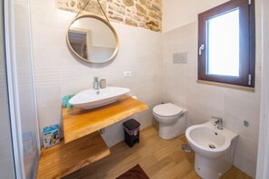 Ein Badezimmer mit einem hölzernen Waschtisch, einem runden Spiegel, einem Bidet und einem Fenster.