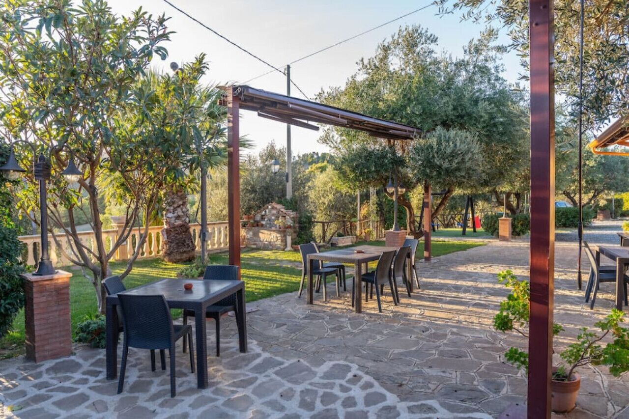 Un patio al aire libre con suelo de piedra, pérgola de metal y mesas con sillas.