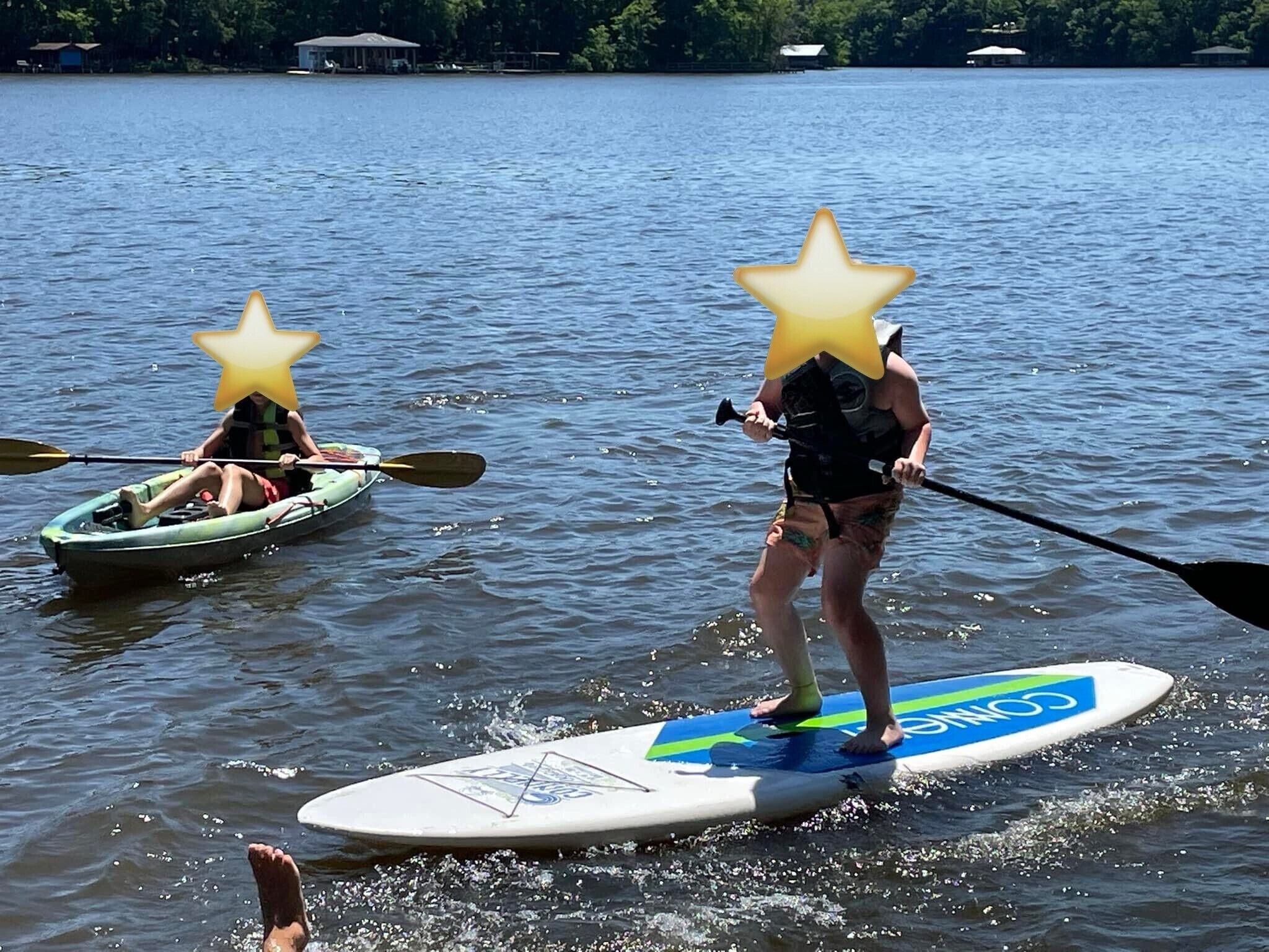 Dos personas en tablas de paddleboard sobre un lago, con un bote pequeño al fondo.