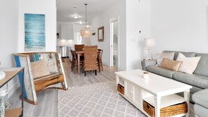 A modern living room with a grey sofa, white coffee table, and a dining area with wicker chairs and a wooden table. - Waterside Retreat (Port St. Joe)