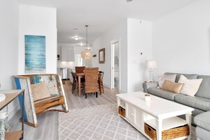 A modern living room with a grey sofa, white coffee table, and a dining area with wicker chairs and a wooden table.