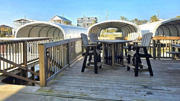 A wooden deck with tables and chairs, a covered walkway, and buildings in the background. - Waterside Retreat (Port St. Joe)