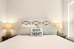 A neatly made bed with a pillow labeled "BEACH THERAPY," two bedside lamps, and a small clock on a bedside table.