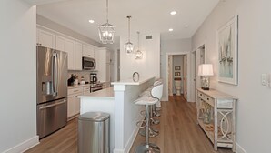A modern kitchen with stainless steel appliances, a central island with bar stools, pendant lighting, and a console table with decorative items. - The Vue 2C (Port St. Joe)