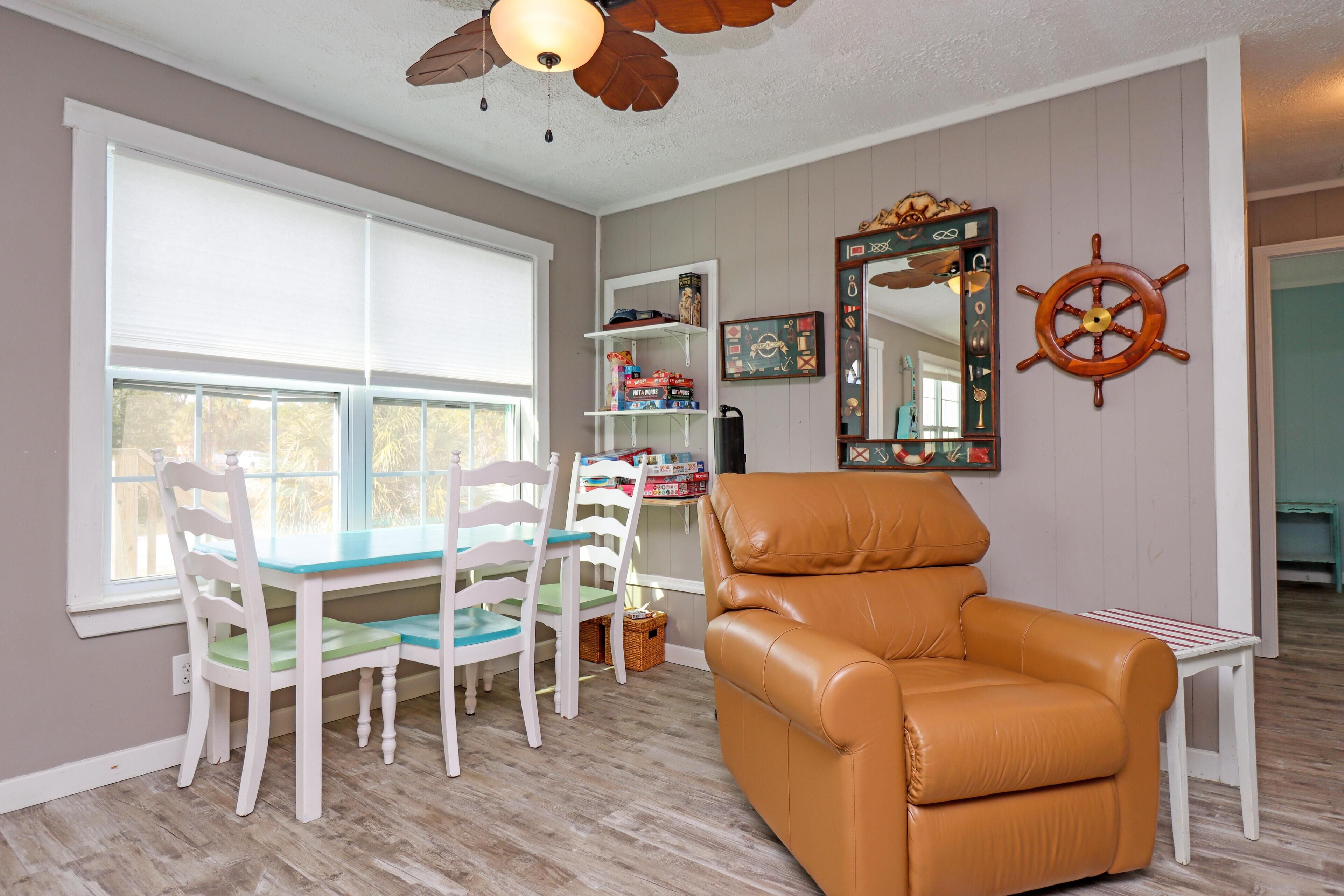 A dining area with a table and chairs, a leather armchair, a ceiling fan, and a wall-mounted mirror with a ship's wheel decoration.