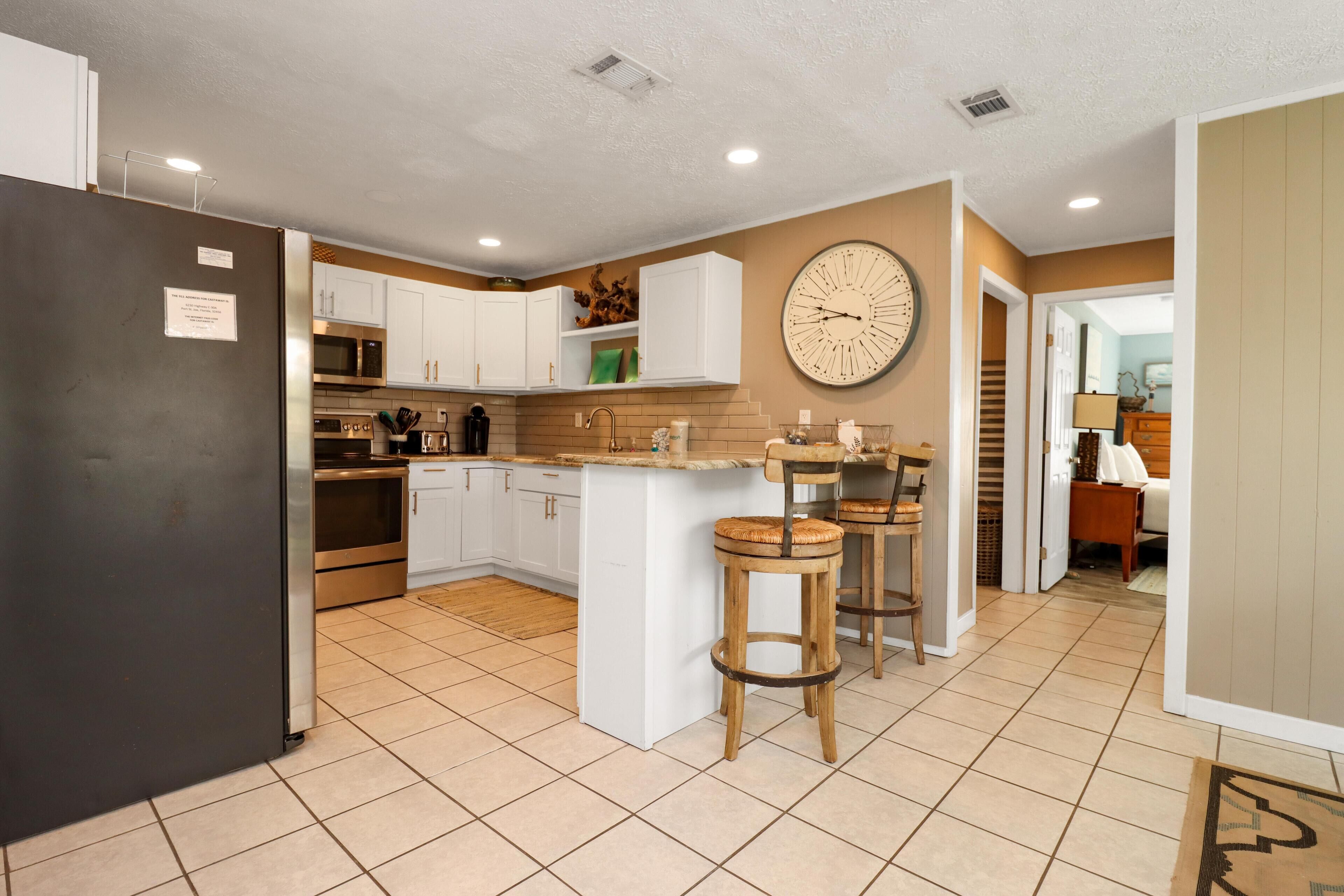 A kitchen with white cabinets, a stainless steel refrigerator, a large clock on the wall, and two wooden stools.