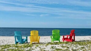 Vier kleurrijke Adirondack-stoelen op rij op een zanderig strand, met de oceaan op de achtergrond.
