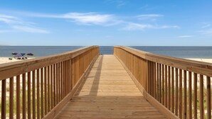 Une promenade en bois menant à une plage où se trouvent des parasols, sous un ciel bleu dégagé.