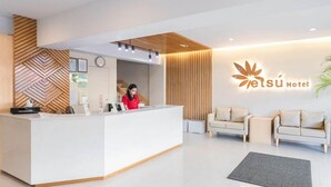 Ein moderner Hotel-Empfangsbereich mit einem weißen Empfangstresen, einer Uhr und einem Logo an der Wand. Im Sitzbereich befinden sich zwei Sofas und ein Blumentopf.