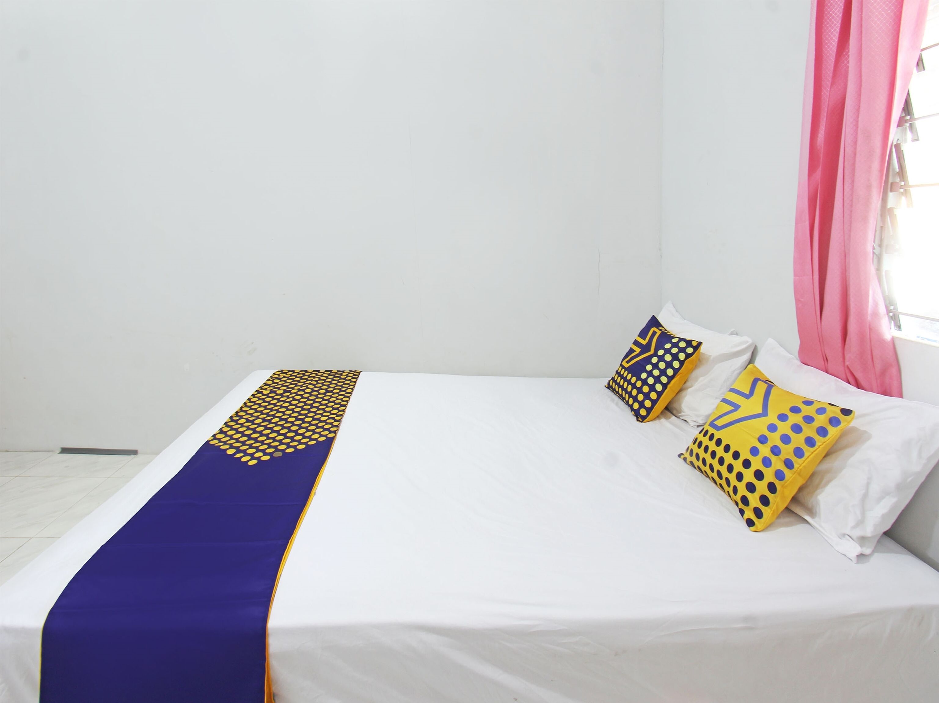 A single bed with a blue and yellow patterned bedspread, white pillows, and a window with pink curtains.