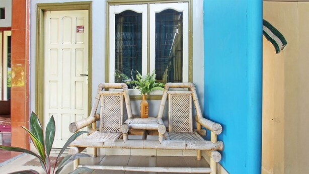A traditional rattan bench with a woven backrest and armrests, placed in front of a building with a blue wall and a yellow door. - OYO Life 92960 Rumah Kos Kita (Gangga)