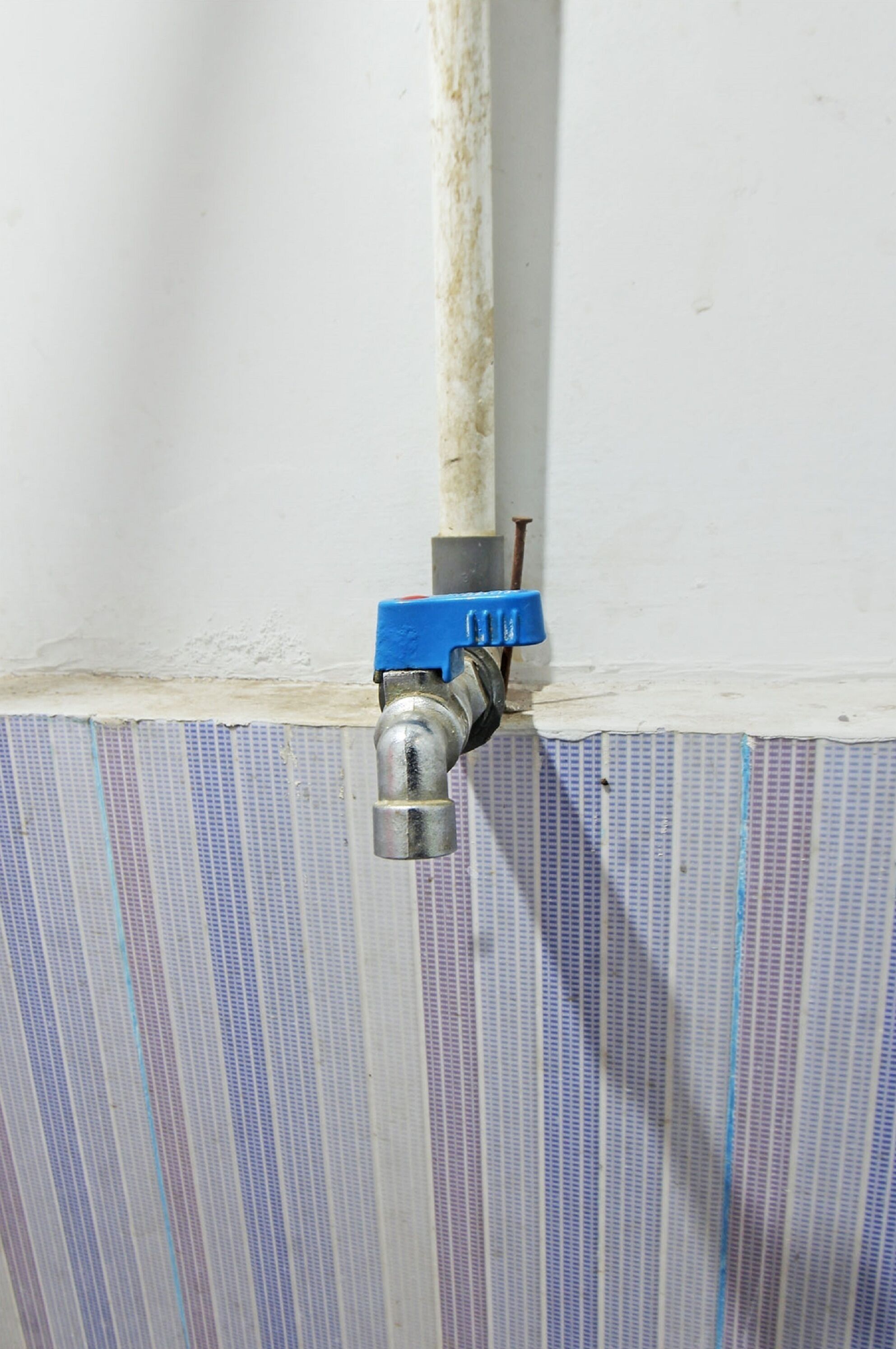 Primer plano de una tubería de metal con una válvula azul, adherida a una pared blanca con signos de desgaste y un patrón de rayas verticales azules.