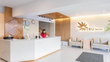 Ein moderner Hotel-Empfangsbereich mit einem weißen Empfangstresen, einer Uhr und einem Logo an der Wand. Im Sitzbereich befinden sich zwei Sofas und ein Blumentopf.