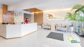 Ein moderner Hotel-Empfangsbereich mit einem weißen Empfangstresen, einer Uhr und einem Logo an der Wand. Im Sitzbereich befinden sich zwei Sofas und ein Blumentopf.