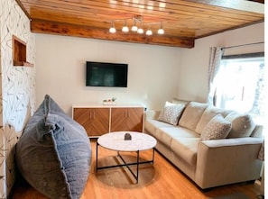 A cozy living room with a sofa, a bean bag, a coffee table, and a TV mounted on the wall. - Aurora Cabin · Walk to Creamer's Field · Fire Pit · Trails · Near UAF (Fairbanks)