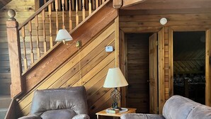 A wooden cabin interior with a staircase, leather sofas, a table, and a lamp. - NEW RIVER GORGE CABINS-ALMOST HEAVEN (Lansing)