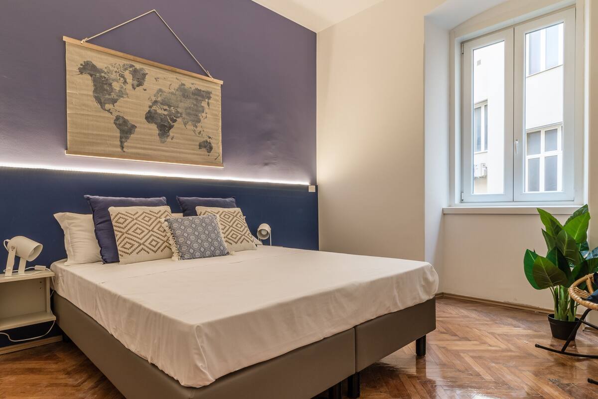 Um quarto com cama, mesa de cabeceira, luminária, mapa-múndi na parede e janela.