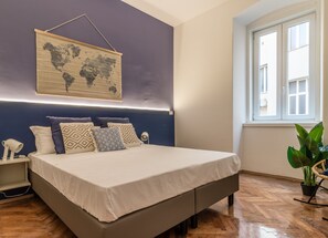 A bedroom with a bed, a nightstand, a lamp, a world map on the wall, and a window.