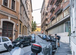 A narrow urban street lined with parked cars and motorcycles, with buildings featuring classic architecture on both sides.