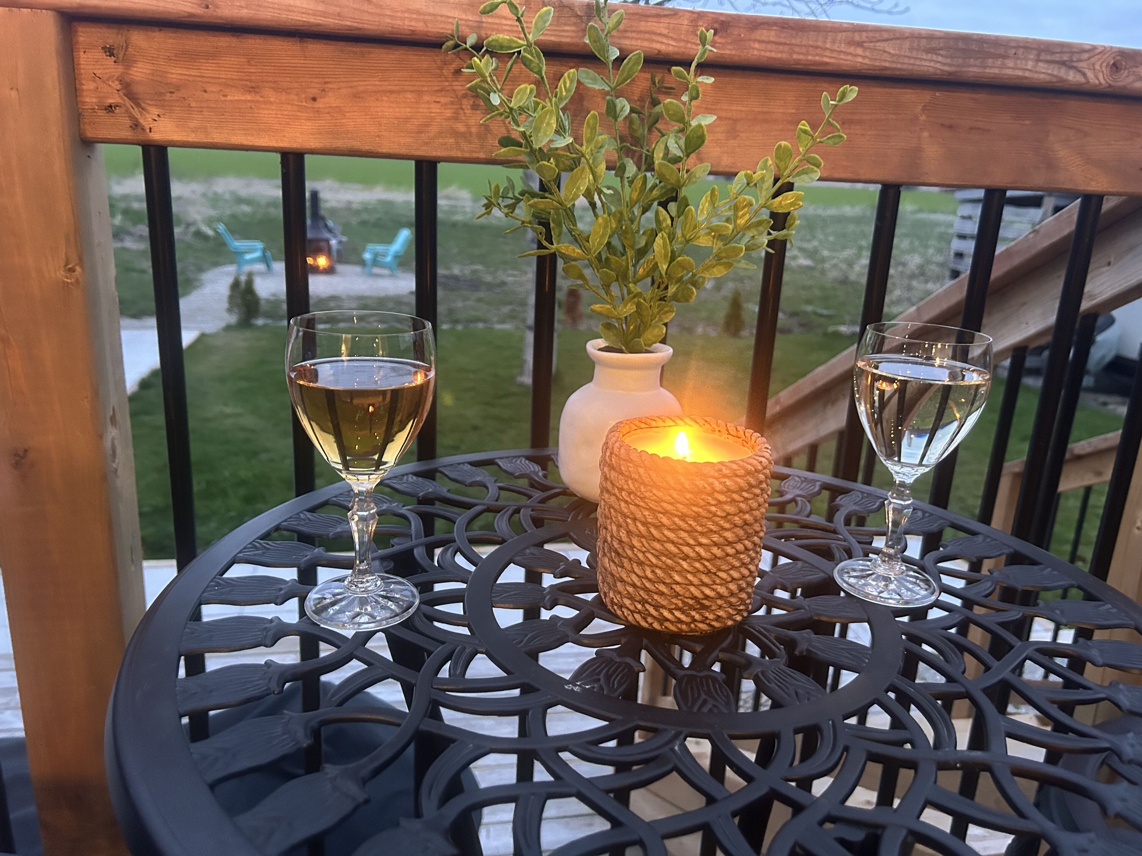 A round metal table with a candle and two glasses of white wine, set on a wooden deck with a view of a grassy area and chairs.