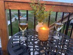 Une table ronde en métal, ornée d’une bougie et de deux verres de vin blanc, est placée sur une terrasse en bois, d’où l’on peut admirer un espace verdoyant et des chaises.