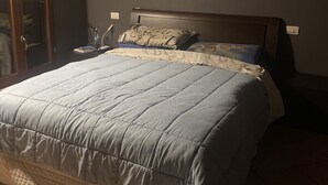 A bedroom with a large bed, a wooden headboard, a nightstand with a lamp, and a window with blinds.