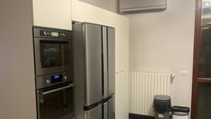 A compact kitchen with a stainless steel refrigerator, oven, and microwave. There is a trash bin and a radiator on the wall.