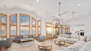 A spacious living room with large windows, a ceiling fan, and a kitchen area visible through an open door. - Oceanfront, Game Room, Decks, Heated Pool, Hot Tub, Ocean Views, Fully Loaded (Corolla)