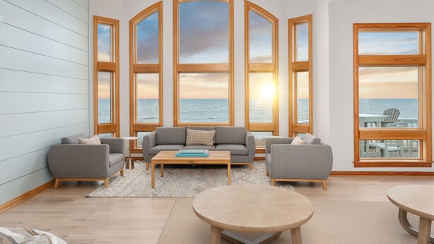 A spacious living room with a large bay window overlooking the sea, a grey sofa, a coffee table, and two round tables. - Oceanfront, Game Room, Decks, Heated Pool, Hot Tub, Ocean Views, Fully Loaded (Corolla)