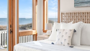 A bedroom with a large bed, white bedding, and a view of the ocean through large windows. - Oceanfront, Game Room, Decks, Heated Pool, Hot Tub, Ocean Views, Fully Loaded (Corolla)