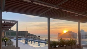 A rooftop terrace with a pool, potted plants, and a view of the sunset over the city skyline. - Hermoso Apto en Santa Marta con Piscina (Gaira)