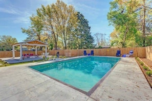 A rectangular swimming pool with a wooden deck surrounding it, a covered seating area with lounge chairs, and a wooden fence in the background.