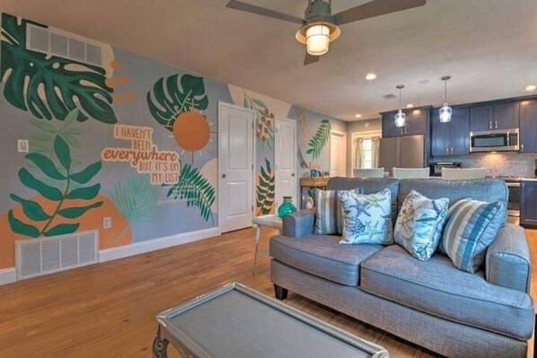 A modern living room with a gray sofa, a coffee table, and a wall mural with tropical leaves and text. - Pensacola Backyard Oasis 
January 2026 Pool Renovation (Gulf Breeze)