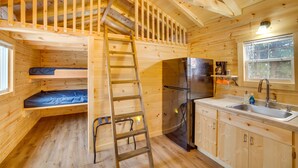 Una cabaña de madera compacta con un altillo para dormir, una cocina con refrigerador y un ventanal que proporciona luz natural.
