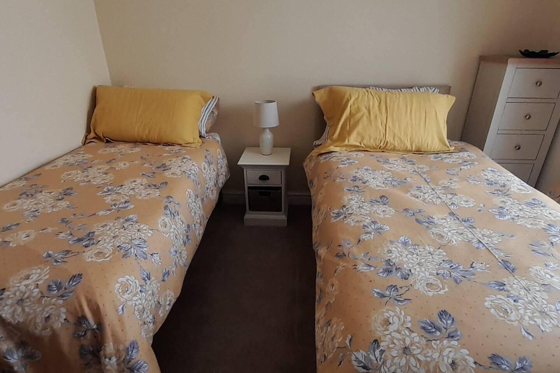 Quarto com duas camas de solteiro, cada uma com colcha florida e travesseiro amarelo. Há uma mesa de cabeceira com abajur entre as camas.