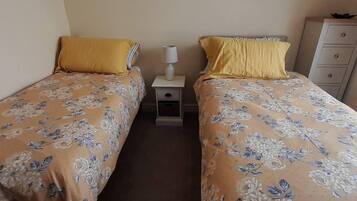 Une chambre avec deux lits simples, chacun recouvert d’une couvre-lit à motifs floraux et agrémenté d’un oreiller jaune. Une table de chevet avec une lampe se trouve entre les deux lits.